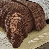 A close-up of a brown quilted blanket draped over a bed with patterned white bedding and a beige rug on a wooden floor.