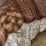 A close-up of a brown quilted velvet blanket draped over a bed with a white and brown floral damask patterned sheet.