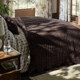 A bed dressed in a dark brown textured quilted comforter and patterned gray and white sheets, styled in a sunlit bedroom with floral curtains and windows.