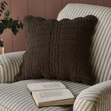 A dark brown quilted throw pillow with scalloped edges resting on a cream and gray striped armchair, with an open book on the seat and a muted pink paneled wall in the background.
