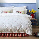 A styled bedroom scene featuring a white duvet cover and matching pillowcases with a delicate multicolor floral and bird print, styled with a red and white checkered bed skirt, a blue paneled headboard, and a red patterned pendant lamp.