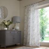White sheer curtains with a delicate light blue and beige floral branch pattern hanging on a window in a styled room with a gray sideboard and lamp.