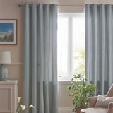 A pair of light blue-gray textured grommet curtains hanging on a window in a styled living room with a beige armchair, potted plant, and wooden floor.