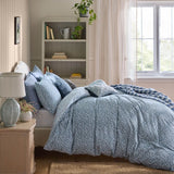 A bed dressed in a light blue and white floral-patterned duvet cover and matching pillowcases, styled in a bright, neutral bedroom with a wooden nightstand, lamp, bookshelf, and window.