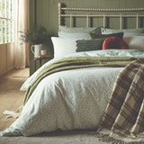 A styled bedroom scene showing a bed dressed in a light sage green patterned duvet cover, layered with an olive green throw blanket and a beige plaid fringed throw. The background features a sage green wooden headboard, beige walls, and a window with soft curtains.